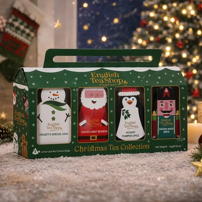 A boxed set of four English Tea Shop Christmas tea bags in a green gift bag with a handle, placed on a snowy surface in front of a blurred Christmas tree and holiday decor.