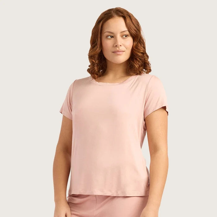 A woman with wavy brown hair wearing a light pink short-sleeve crewneck top and matching pink bottoms, photographed against a plain light gray studio background.