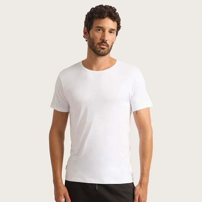 A man wearing a plain white short-sleeve crewneck t-shirt, paired with black pants, standing against a solid light beige background.