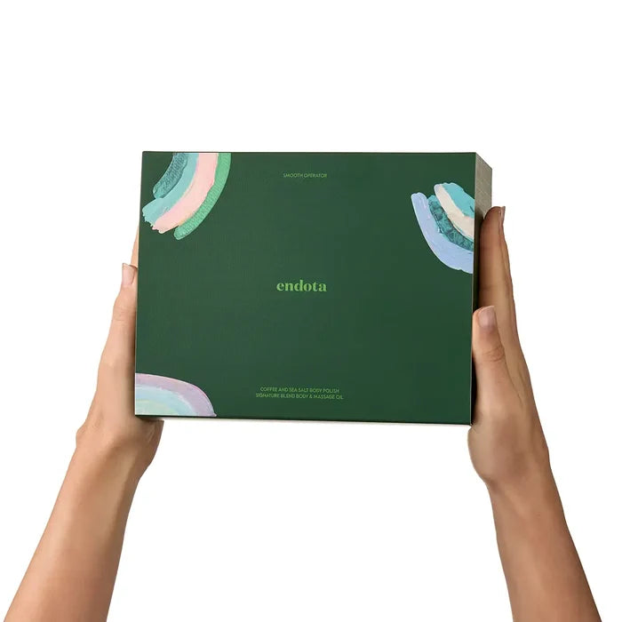 A person's hands holding a dark green Endota skincare box against a solid white background. The box features abstract pastel rainbow brushstrokes in the corners and the brand name 'endota' printed in the center.