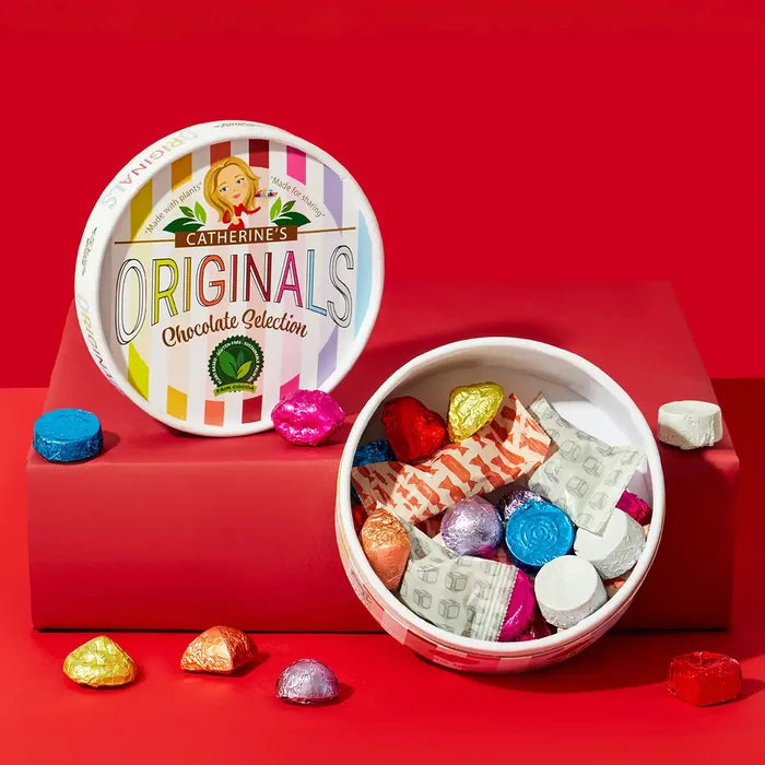 Catherine's Originals chocolate selection in a round tin, shown open with assorted foil-wrapped chocolates and candy pieces spilling out, next to the branded lid on a red background.